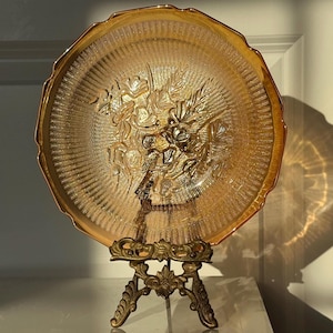 Puede incluir: Un plato de vidrio ámbar vintage con un diseño floral, exhibido en un soporte de metal dorado.
