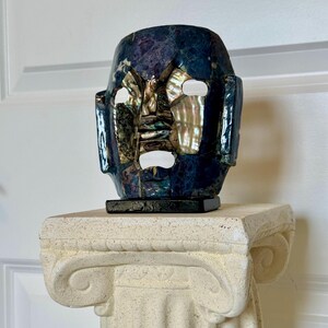May include: A decorative, abstract face sculpture with a dark blue, iridescent finish. The face features inlaid iridescent shell accents for the eyes, nose, and mouth. The sculpture sits on a small black base, displayed on a white, ornate pedestal.