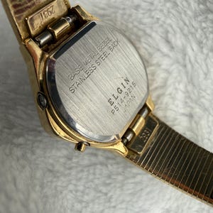 May include: Close-up of a gold-toned Elgin watch. The watch features a rectangular face with a stainless steel back and a base metal bezel. The watch band is a gold-toned, flexible mesh style. The watch is marked "Japan" and "2931".