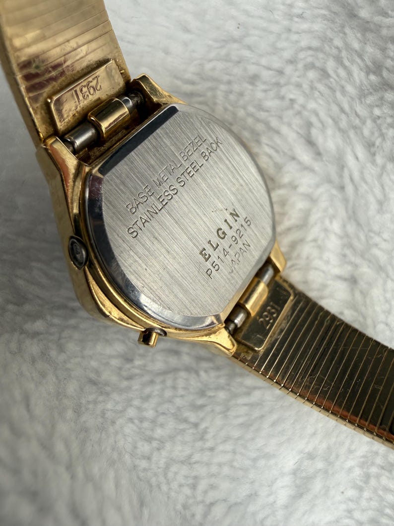 May include: Close-up of a gold-toned Elgin watch. The watch features a rectangular face with a stainless steel back and a base metal bezel. The watch band is a gold-toned, flexible mesh style. The watch is marked "Japan" and "2931".