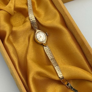May include: A gold-tone analog wristwatch featuring a small, oval-shaped face with a white dial and gold markers. The watch has a textured, link-style metal band with a hammered finish, providing a distinct visual pattern along the entire length of the strap.