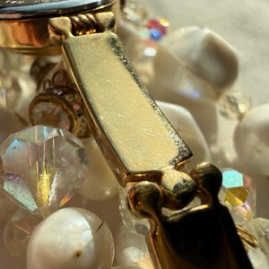 May include: Close-up of a vintage necklace featuring a gold-tone clasp and a mix of white, iridescent, and clear beads. The beads vary in shape and size, creating a layered effect. The clasp is rectangular and has a textured surface.