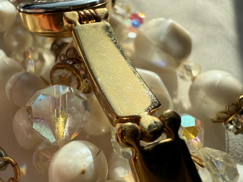 May include: Close-up of a vintage necklace featuring a gold-tone clasp and a mix of white, iridescent, and clear beads. The beads vary in shape and size, creating a layered effect. The clasp is rectangular and has a textured surface.