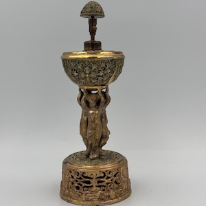 Pode incluir: Um queimador de incenso antigo em latão com intrincados filigranas e figuras de querubins. O queimador apresenta um topo em forma de cúpula, um centro em forma de tigela e uma base detalhada. A cor geral é uma mistura de ouro e bronze, com detalhes ornamentados.