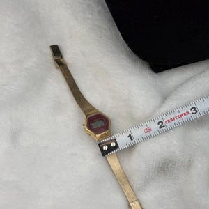 May include: A vintage gold-toned digital watch with a rectangular red display. The watch band is a thin, gold mesh. The watch is measured with a Craftsman tape measure. The watch band is approximately 0.75 inches wide.