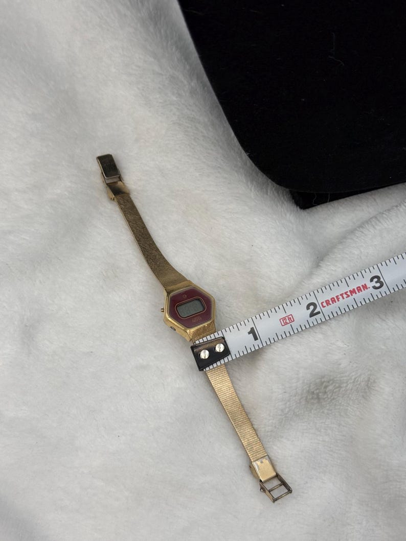 May include: A vintage gold-toned digital watch with a rectangular red display. The watch band is a thin, gold mesh. The watch is measured with a Craftsman tape measure. The watch band is approximately 0.75 inches wide.