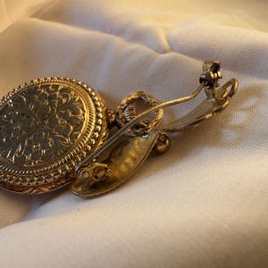 May include: A vintage gold-toned brooch with an oval, floral-patterned pendant. The brooch has a pin closure on the back. The pendant is attached to the pin with a small loop. The brooch is set against a white fabric.