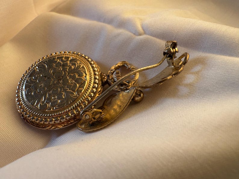 May include: A vintage gold-toned brooch with an oval, floral-patterned pendant. The brooch has a pin closure on the back. The pendant is attached to the pin with a small loop. The brooch is set against a white fabric.