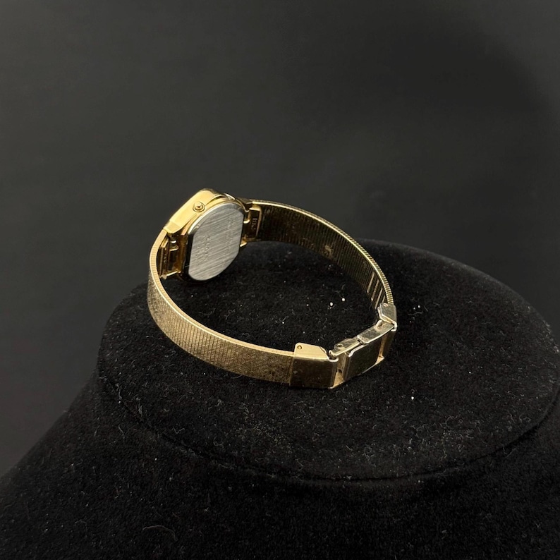 May include: A gold-toned wristwatch with a rectangular face and a textured band. The watch is displayed on a black surface, showcasing its elegant design. The watch band appears to be approximately 0.5 inches wide.