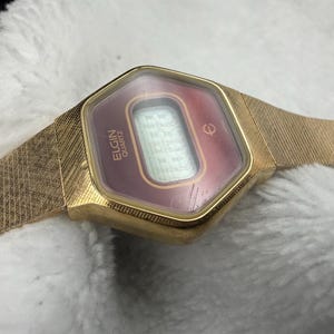 May include: A vintage Elgin Quartz watch with a gold-tone case and band. The watch face is a burgundy color with a digital display. The band has a textured, woven design. The watch is displayed on a white, textured surface.