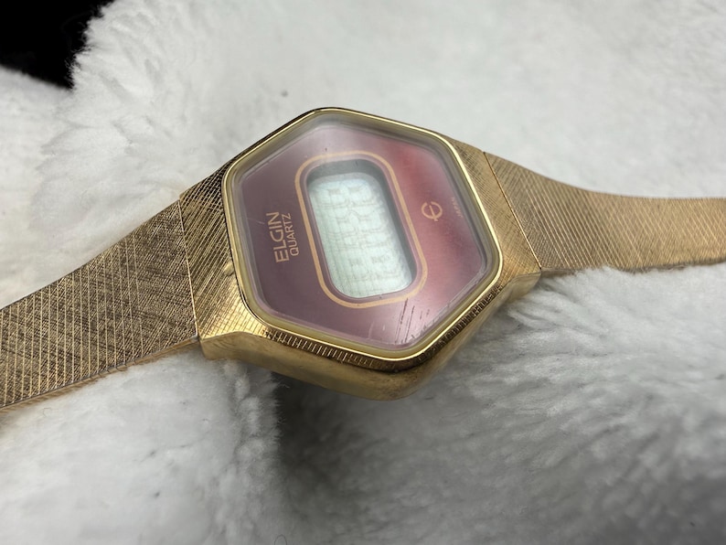 May include: A vintage Elgin Quartz watch with a gold-tone case and band. The watch face is a burgundy color with a digital display. The band has a textured, woven design. The watch is displayed on a white, textured surface.