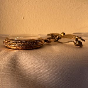 May include: A vintage gold-toned compact with a clear lid and ornate filigree detailing. The compact is connected to a gold-toned clip with a decorative butterfly design. The compact is resting on a white surface.