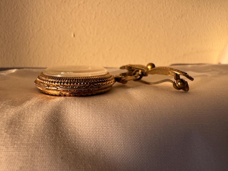 May include: A vintage gold-toned compact with a clear lid and ornate filigree detailing. The compact is connected to a gold-toned clip with a decorative butterfly design. The compact is resting on a white surface.