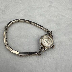May include: A silver-toned vintage wristwatch with a rectangular link band. The watch face is round with a white background and black numerals. The watch has decorative heart-shaped accents on the sides.