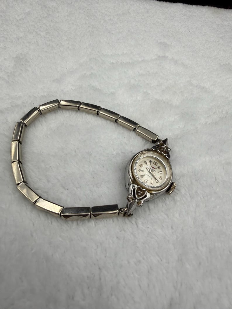 May include: A silver-toned vintage wristwatch with a rectangular link band. The watch face is round with a white background and black numerals. The watch has decorative heart-shaped accents on the sides.