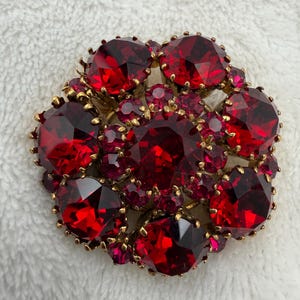 May include: A circular brooch featuring a cluster of faceted deep red stones set in a gold-toned metal frame. The design consists of several large, round, brilliant-cut red crystals surrounded by smaller red accent stones, all held by metal prongs.
