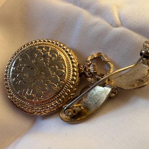 May include: A gold-toned vintage brooch with a round, floral-patterned face and a decorative border. The brooch has a pin closure on the back. The jewelry is set on a white fabric background.