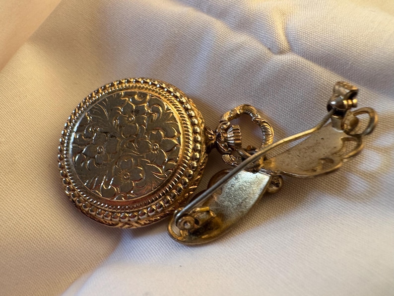 May include: A gold-toned vintage brooch with a round, floral-patterned face and a decorative border. The brooch has a pin closure on the back. The jewelry is set on a white fabric background.