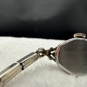 May include: Close-up of a vintage silver-toned wristwatch. The watch features a rectangular face and a delicate, articulated band. The band is marked "MIRA-FLEX" and "20K G.F. US". The watch is set against a soft, white background.