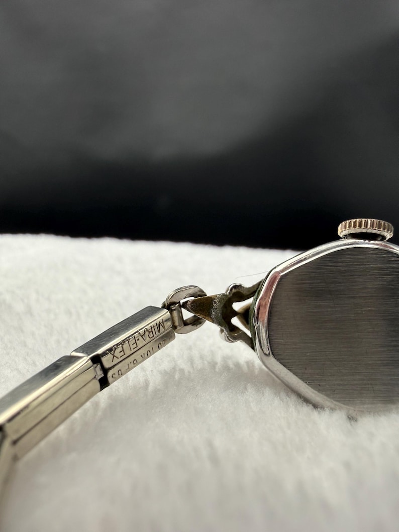 May include: Close-up of a vintage silver-toned wristwatch. The watch features a rectangular face and a delicate, articulated band. The band is marked "MIRA-FLEX" and "20K G.F. US". The watch is set against a soft, white background.
