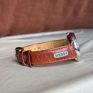 Puede incluir: Una correa de reloj de cuero marr&oacute;n con una esfera plateada. La correa tiene un cierre de hebilla y un peque&ntilde;o parche rectangular con la palabra "FOSSIL" y el n&uacute;mero "315503" en azul. El reloj est&aacute; sobre un fondo neutro.