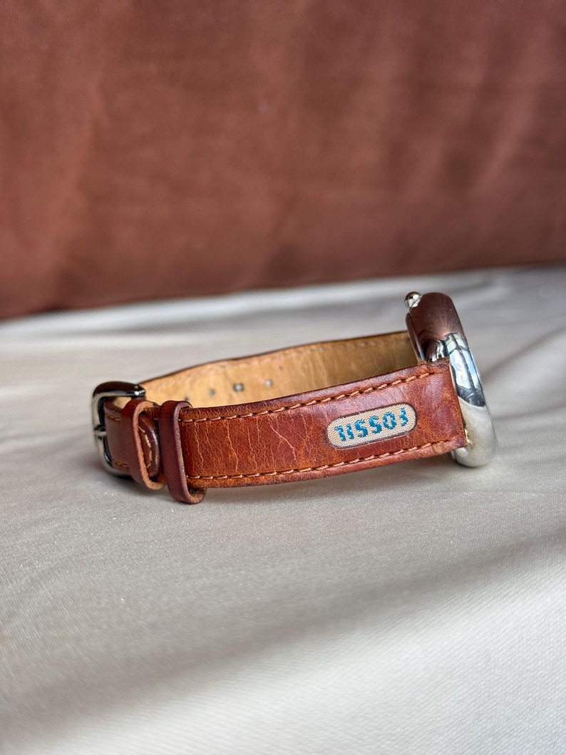 Puede incluir: Una correa de reloj de cuero marr&oacute;n con una esfera plateada. La correa tiene un cierre de hebilla y un peque&ntilde;o parche rectangular con la palabra "FOSSIL" y el n&uacute;mero "315503" en azul. El reloj est&aacute; sobre un fondo neutro.