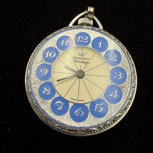 May include: A vintage pocket watch with a silver-tone case and a blue and cream face. The watch face features blue numerals and the words "American Heritage" and "Swiss Made". The watch has a loop at the top for attaching a chain.