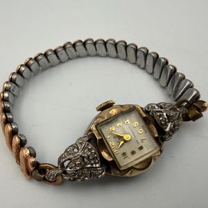 May include: A vintage wristwatch with a square face, gold-tone hands, and numerals. The watch features an ornate, silver-tone metal casing with decorative accents and a flexible, two-tone expansion band made of silver and copper-colored metal links.