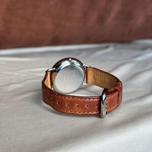 Puede incluir: Una correa de reloj de cuero marr&oacute;n con una hebilla plateada est&aacute; sujeta a una esfera plateada. La esfera del reloj es redonda y la correa es de cuero marr&oacute;n. La palabra "FOSSIL" est&aacute; grabada en el interior de la correa.