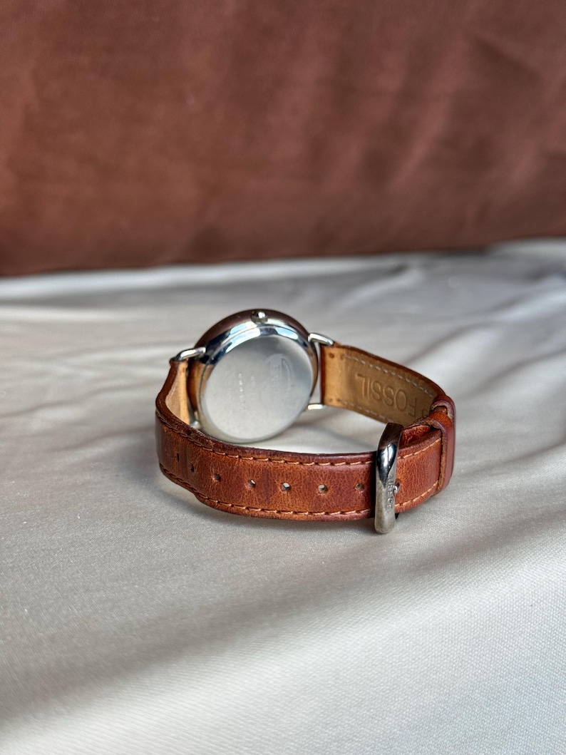 Puede incluir: Una correa de reloj de cuero marr&oacute;n con una hebilla plateada est&aacute; sujeta a una esfera plateada. La esfera del reloj es redonda y la correa es de cuero marr&oacute;n. La palabra "FOSSIL" est&aacute; grabada en el interior de la correa.