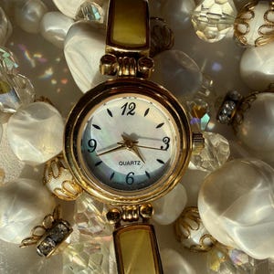 May include: A gold-toned quartz watch with a mother-of-pearl face, featuring black numerals and hands. The watch has a rectangular band with a light-colored inlay. The watch is surrounded by white beads and crystal accents.