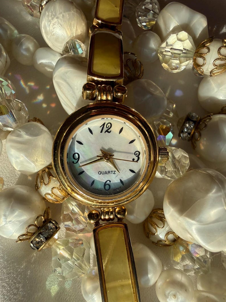 May include: A gold-toned quartz watch with a mother-of-pearl face, featuring black numerals and hands. The watch has a rectangular band with a light-colored inlay. The watch is surrounded by white beads and crystal accents.