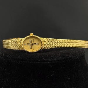 May include: A gold-toned wristwatch with a woven band and a round face. The watch face features a cross design with small gemstones and a small diamond. The watch is displayed on a black surface.