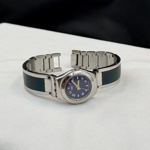May include: A wristwatch featuring a round dark blue dial with gold-tone hands and white numerals. The watch has a silver-tone metal link bracelet with dark green rectangular insets integrated into the band, creating a sleek and polished look.