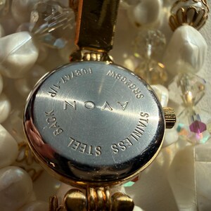 May include: Close-up of an Avon watch with a stainless steel back. The watch is gold-toned and surrounded by a beaded necklace with white and clear beads. The watch face has the text "AVON" and "STAINLESS STEEL".