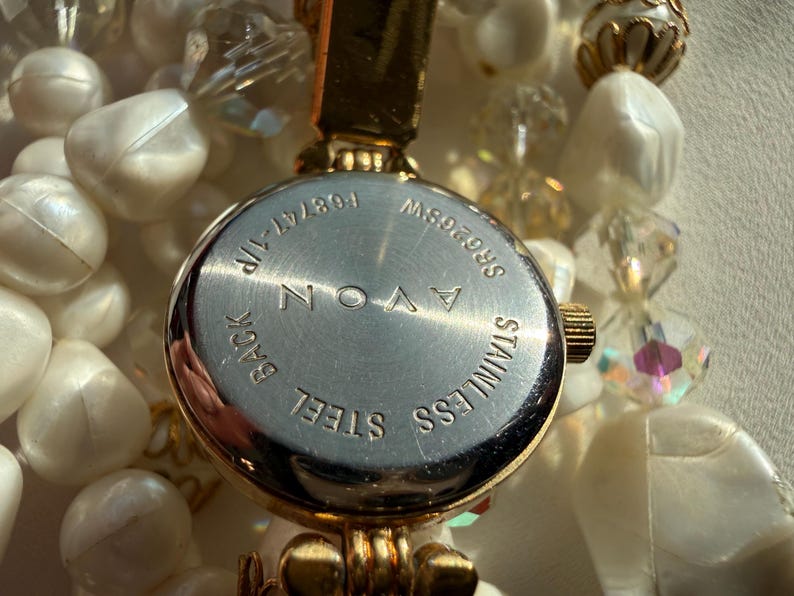 May include: Close-up of an Avon watch with a stainless steel back. The watch is gold-toned and surrounded by a beaded necklace with white and clear beads. The watch face has the text "AVON" and "STAINLESS STEEL".