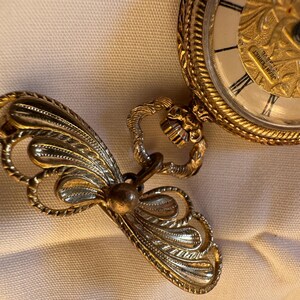 May include: A gold-toned pocket watch with an ornate design and Roman numeral hour markers. The watch is connected to a decorative, stylized butterfly-shaped fob. The watch face has the text "JEAN PERRET".