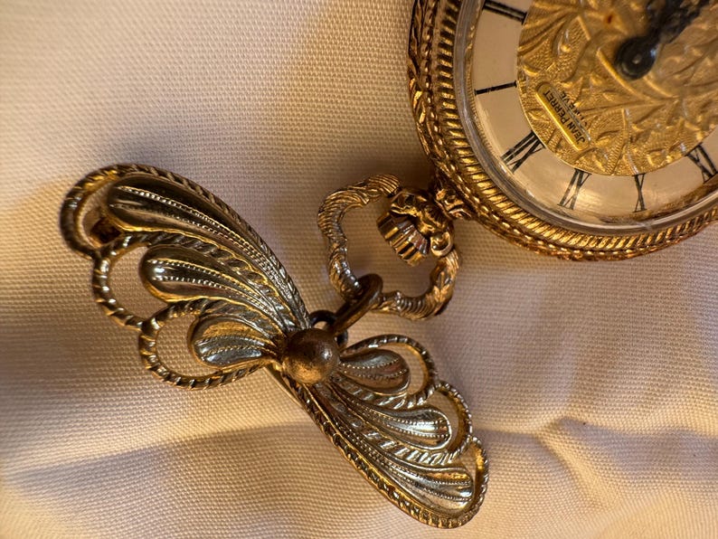 May include: A gold-toned pocket watch with an ornate design and Roman numeral hour markers. The watch is connected to a decorative, stylized butterfly-shaped fob. The watch face has the text "JEAN PERRET".