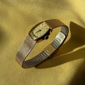 May include: A vintage gold-toned wristwatch with a mesh band. The watch face is rectangular with a cream-colored dial and black hands. The watch is set against a yellow-gold background.