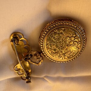 May include: A vintage gold-toned brooch with a round, floral-patterned pendant and a decorative pin. The pendant is connected to the pin with a small chain. The brooch is likely made of metal and features intricate detailing.