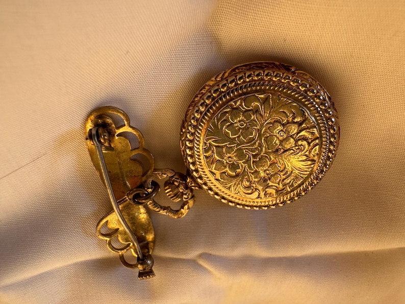 May include: A vintage gold-toned brooch with a round, floral-patterned pendant and a decorative pin. The pendant is connected to the pin with a small chain. The brooch is likely made of metal and features intricate detailing.