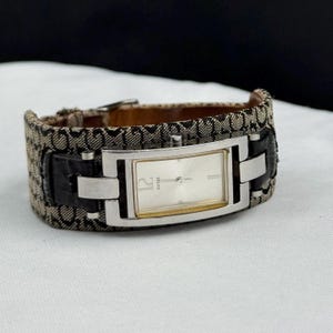 May include: A rectangular Guess watch with a silver face and a brown and black patterned band. The watch has a silver bezel and black leather accents. The watch face displays the number 12. The watch is on a white surface.