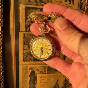 May include: A gold-toned pocket watch brooch with a decorative bow and a small, round watch face. The watch face has Roman numerals and ornate detailing. The brooch is held against a backdrop of vintage newspaper clippings.