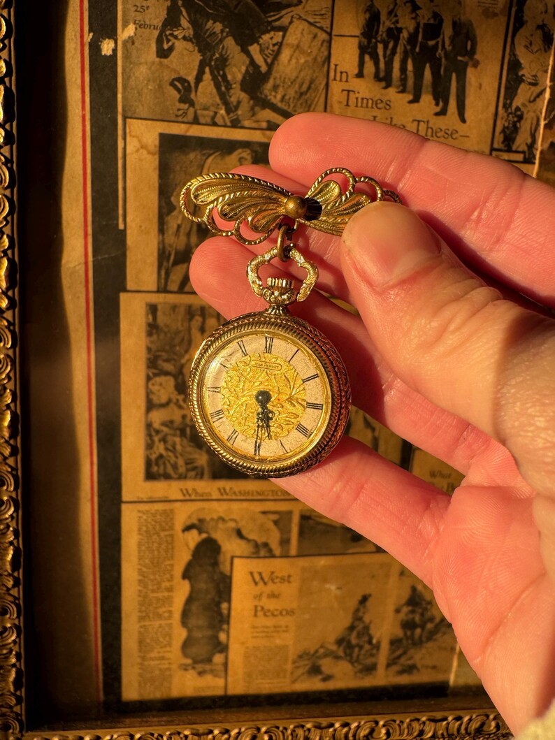 May include: A gold-toned pocket watch brooch with a decorative bow and a small, round watch face. The watch face has Roman numerals and ornate detailing. The brooch is held against a backdrop of vintage newspaper clippings.