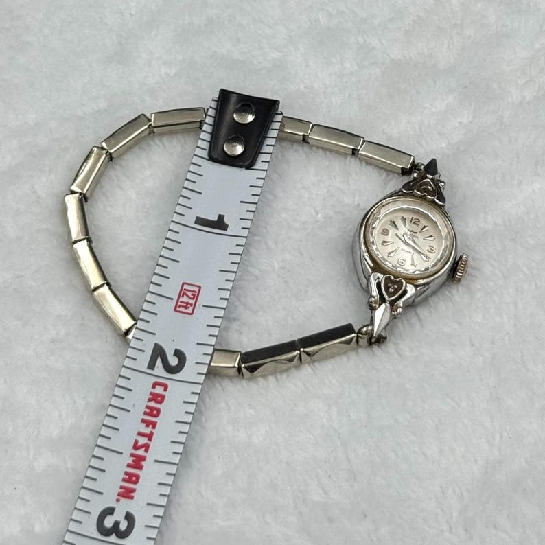 May include: A vintage silver-toned wristwatch with a white face and decorative accents. The watch is measured with a Craftsman ruler, showing a length of approximately 2 inches. The watch band is made of small, rectangular links.