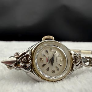 May include: A vintage Waltham wristwatch with a silver-tone case and a white dial. The watch features a gold-tone crown and decorative floral accents on the sides. The watch face displays the numbers 3, 6, 9, and 12.