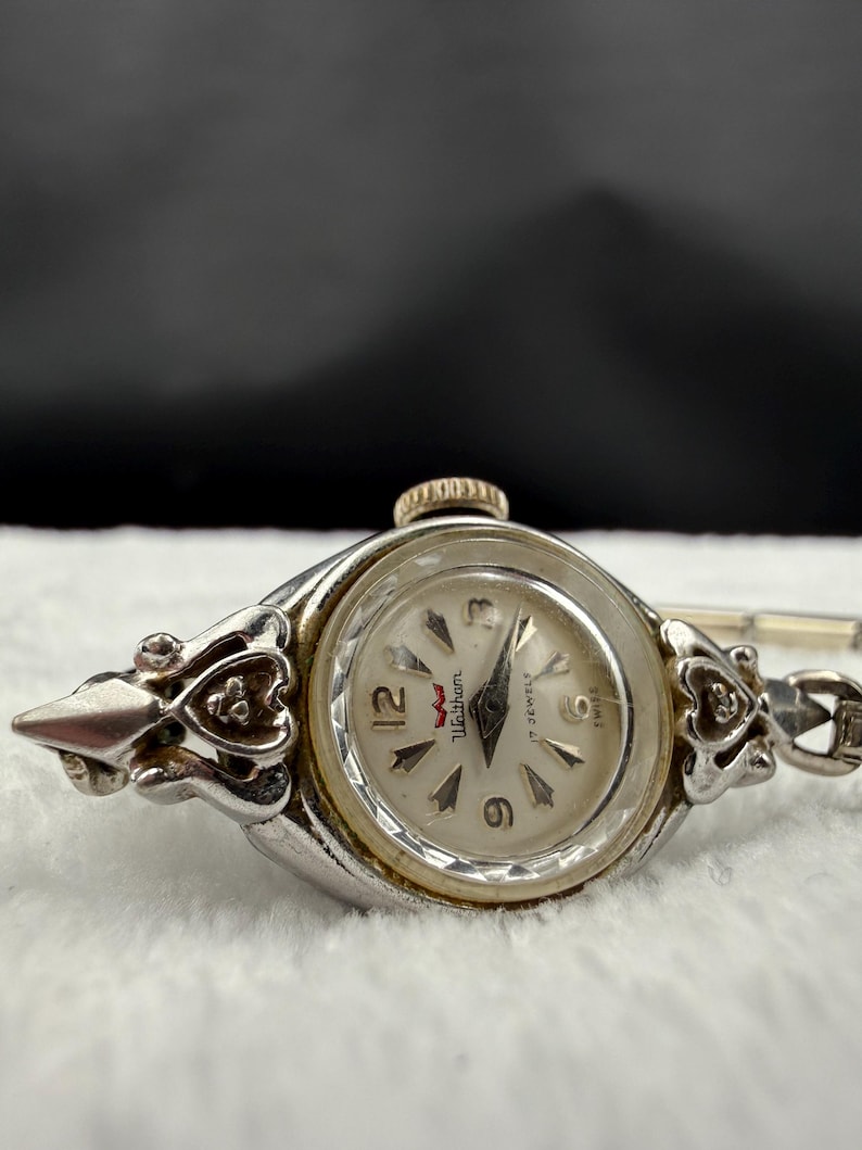 May include: A vintage Waltham wristwatch with a silver-tone case and a white dial. The watch features a gold-tone crown and decorative floral accents on the sides. The watch face displays the numbers 3, 6, 9, and 12.