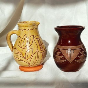 May include: Two ceramic vases. One is a small pitcher with a handle, tan with brown leaf and geometric designs, and an orange base. The other is a dark red vase with a geometric pattern in a lighter brown color.