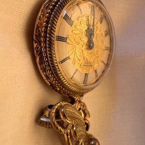 May include: A vintage gold-toned pocket watch with a detailed floral design on the face. The watch has black Roman numerals and ornate detailing around the bezel and chain attachment. The watch hands point to approximately 12:00.