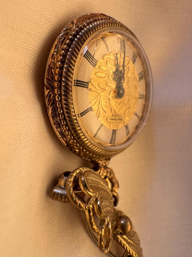 May include: A vintage gold-toned pocket watch with a detailed floral design on the face. The watch has black Roman numerals and ornate detailing around the bezel and chain attachment. The watch hands point to approximately 12:00.